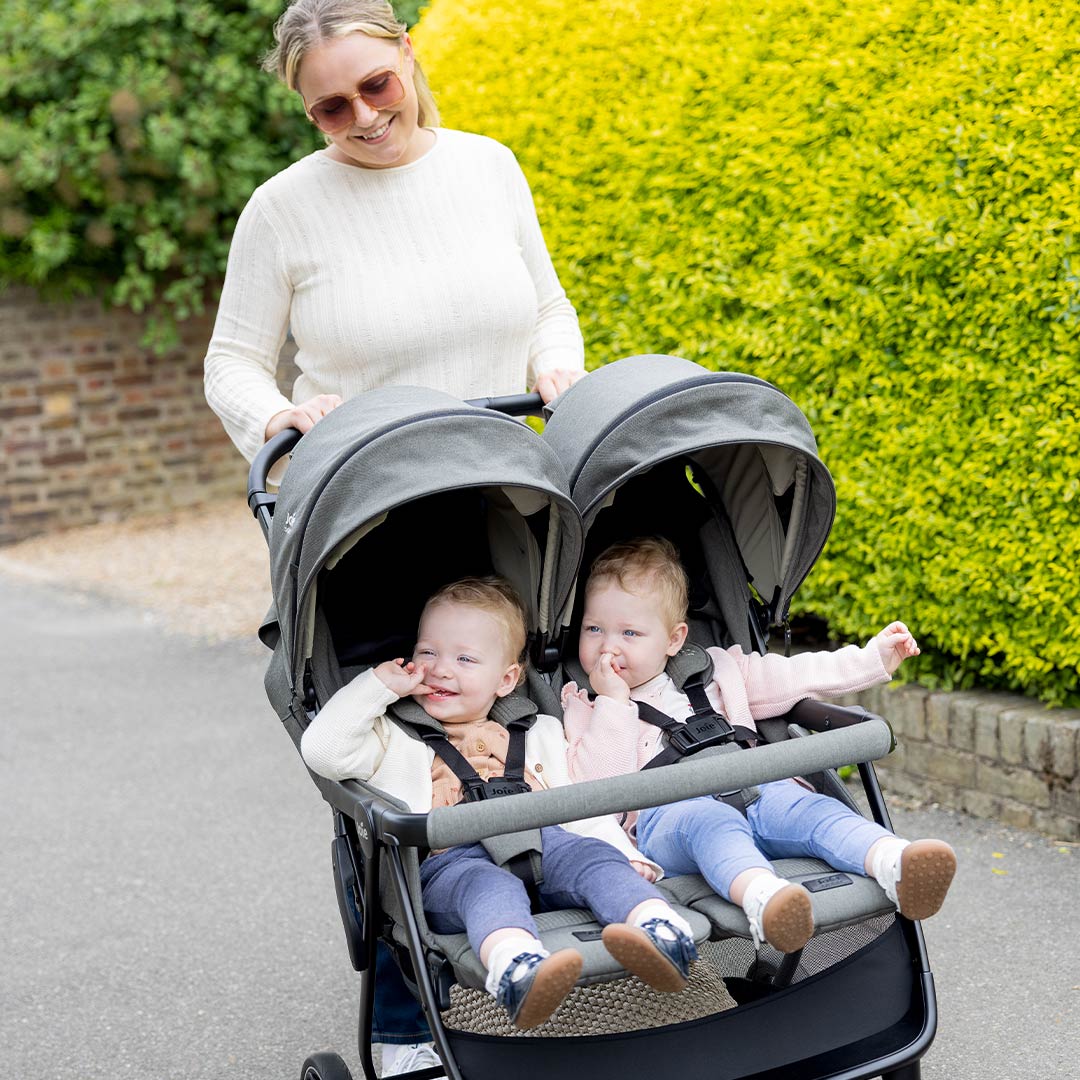 Smiling mother pushing her twins in a grey Joie side-by-side double stroller along a sunny lane beside a bright green hedge