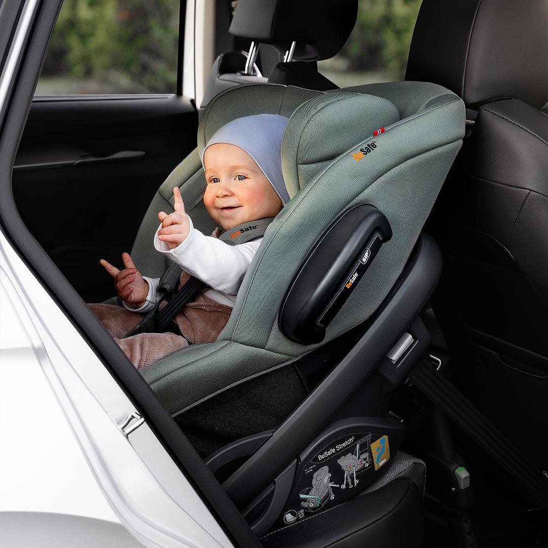 Smiling baby waving from a sage green BeSafe rotating car seat in the back of a car