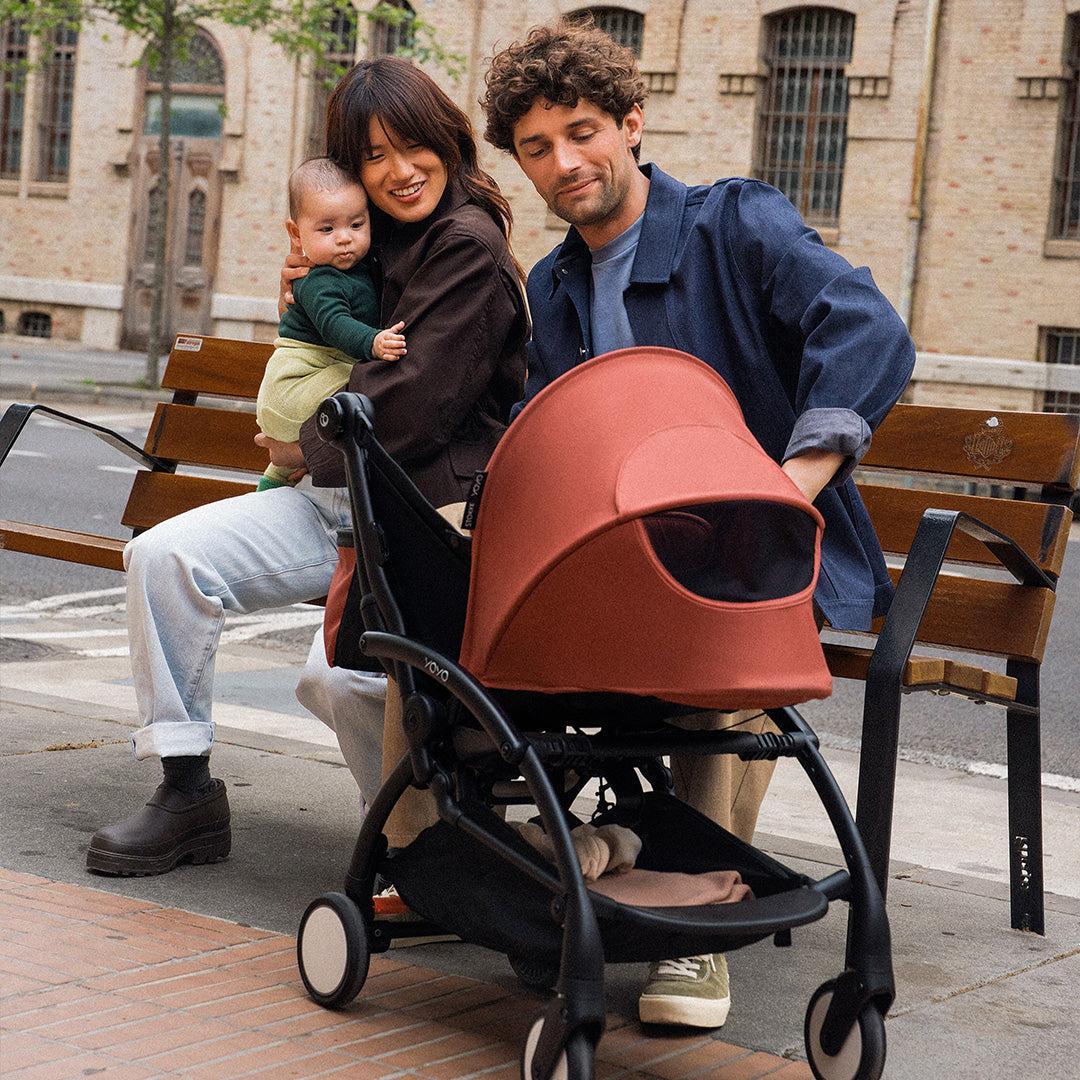 Smiling parents on a city bench, mum cuddling their baby, beside a terracotta Stokke YOYO stroller