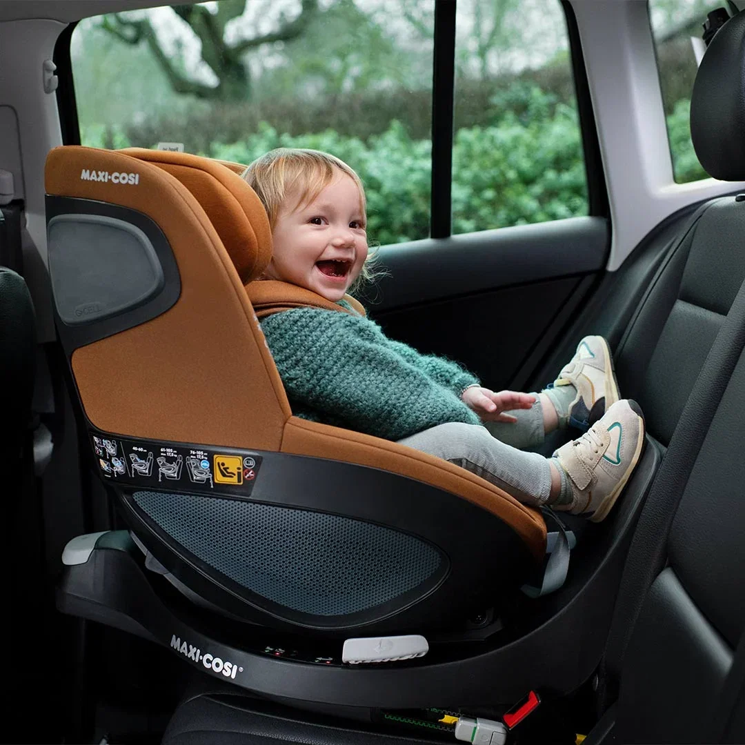Laughing toddler in a green knitted jumper sitting in a cognac Maxi-Cosi rotating car seat in the back of a car
