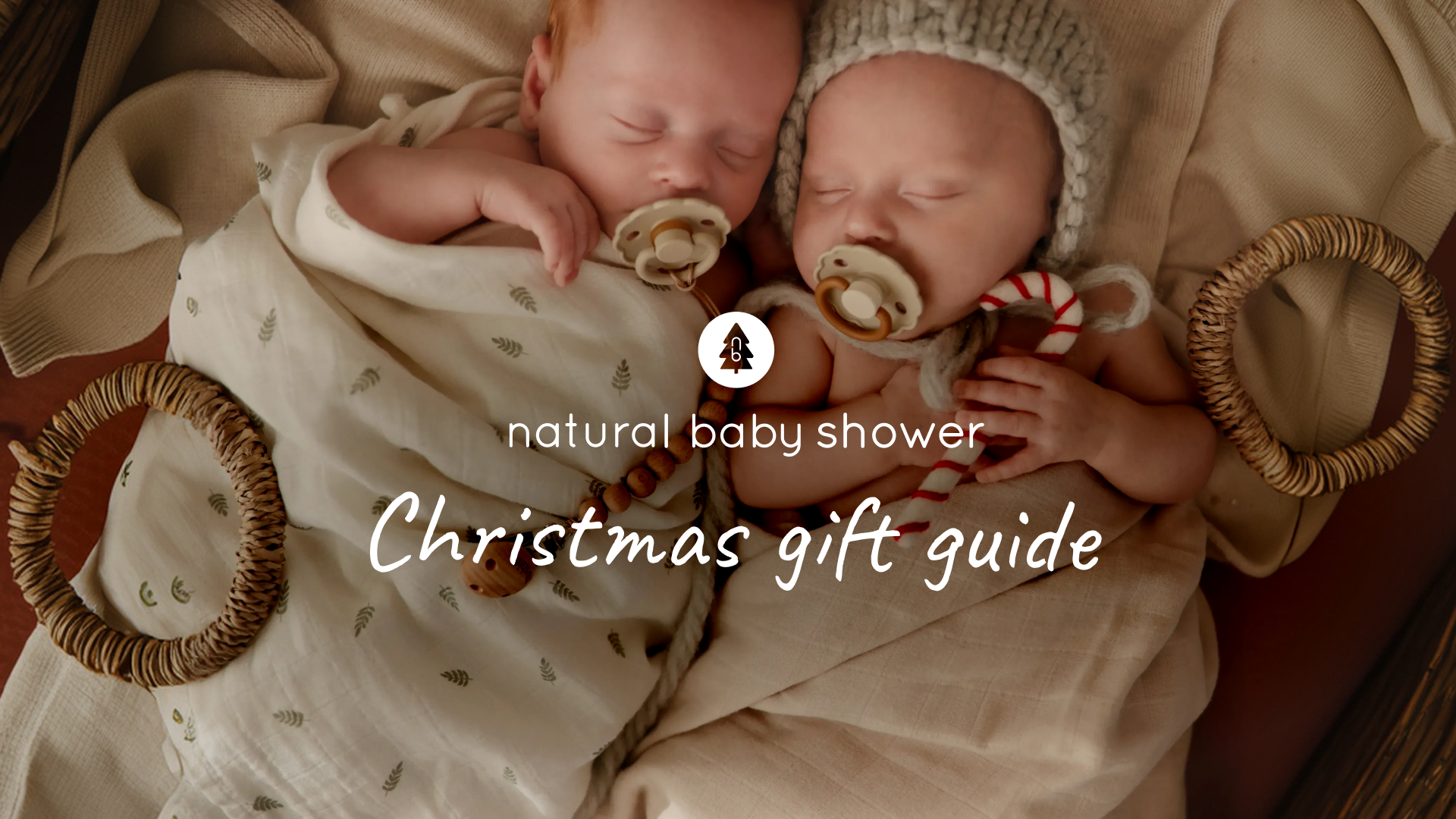 Two newborn babies sleeping side by side in cream muslin wraps, one holding a candy cane, in a woven basket