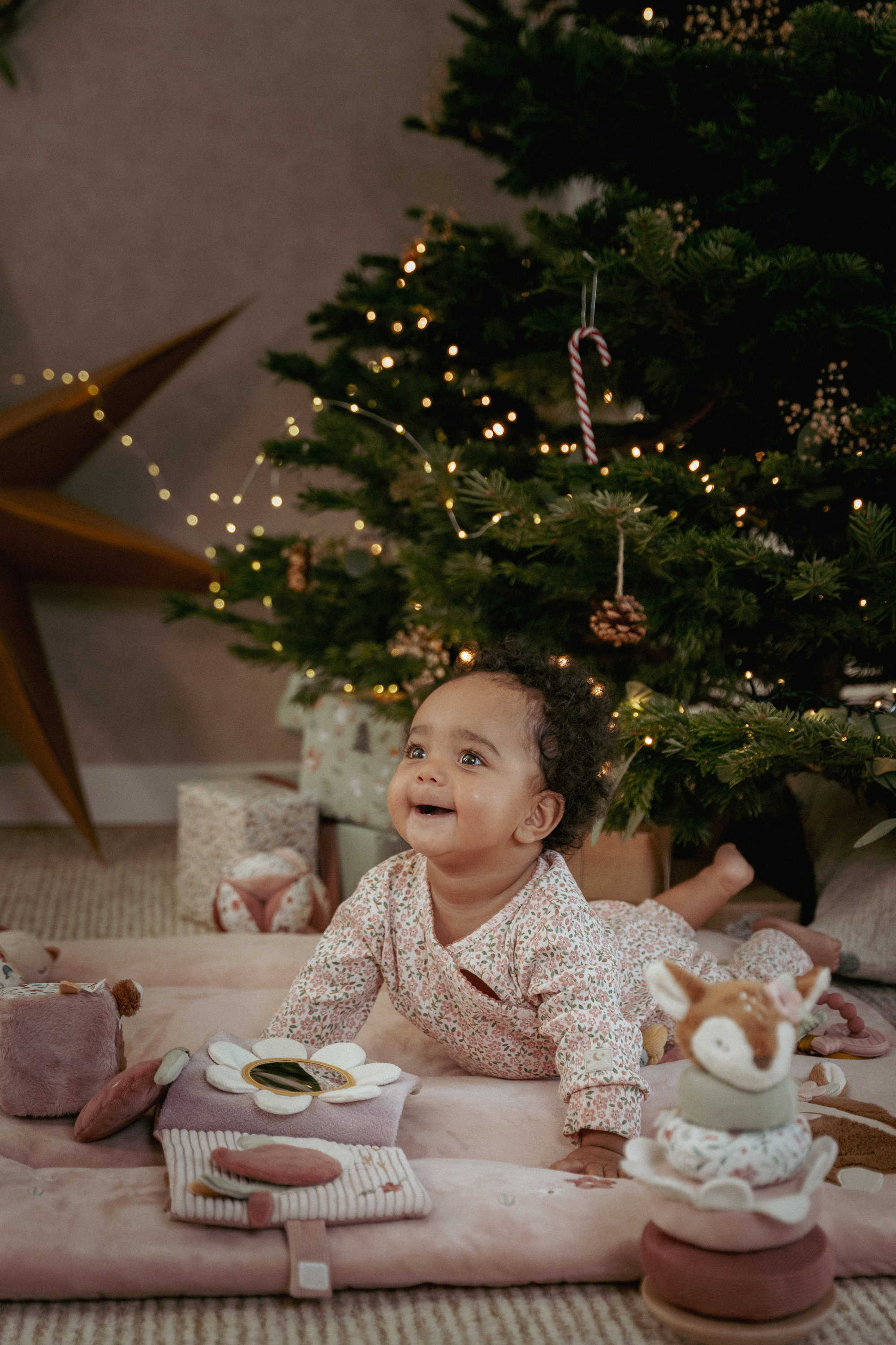 Smiling baby in a floral sleepsuit on a soft pink play mat under a lit Christmas tree, surrounded by a fawn stacking toy, soft activity toys and wrapped gifts