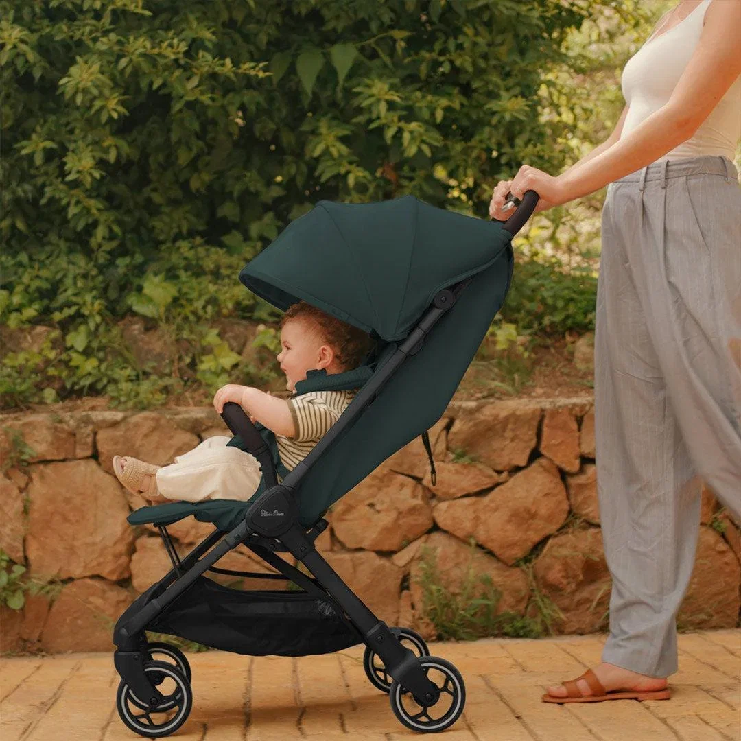 Parent pushing a smiling baby in a teal Silver Cross compact stroller along a sunny garden path beside a stone wall