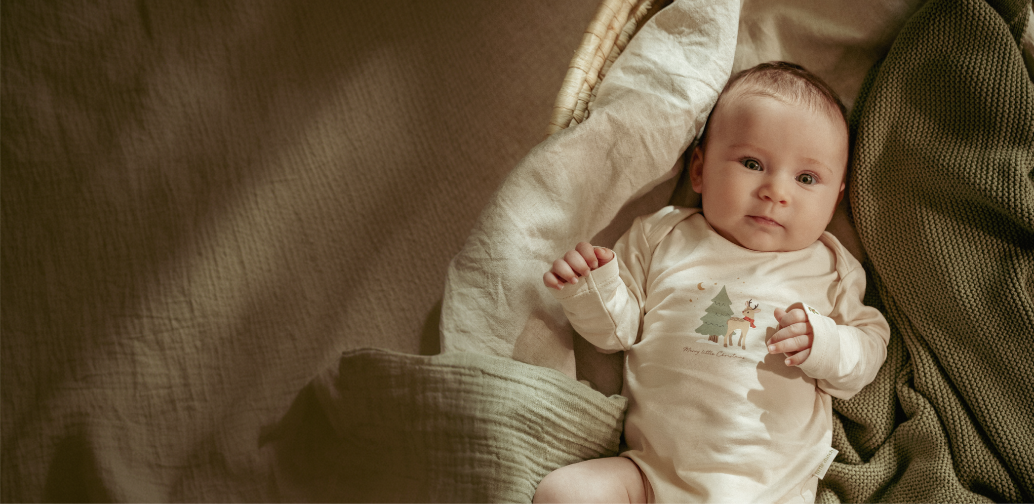 Baby in a cream Merry Little Christmas reindeer bodysuit lying in a woven Moses basket on soft muslin and an olive knitted blanket