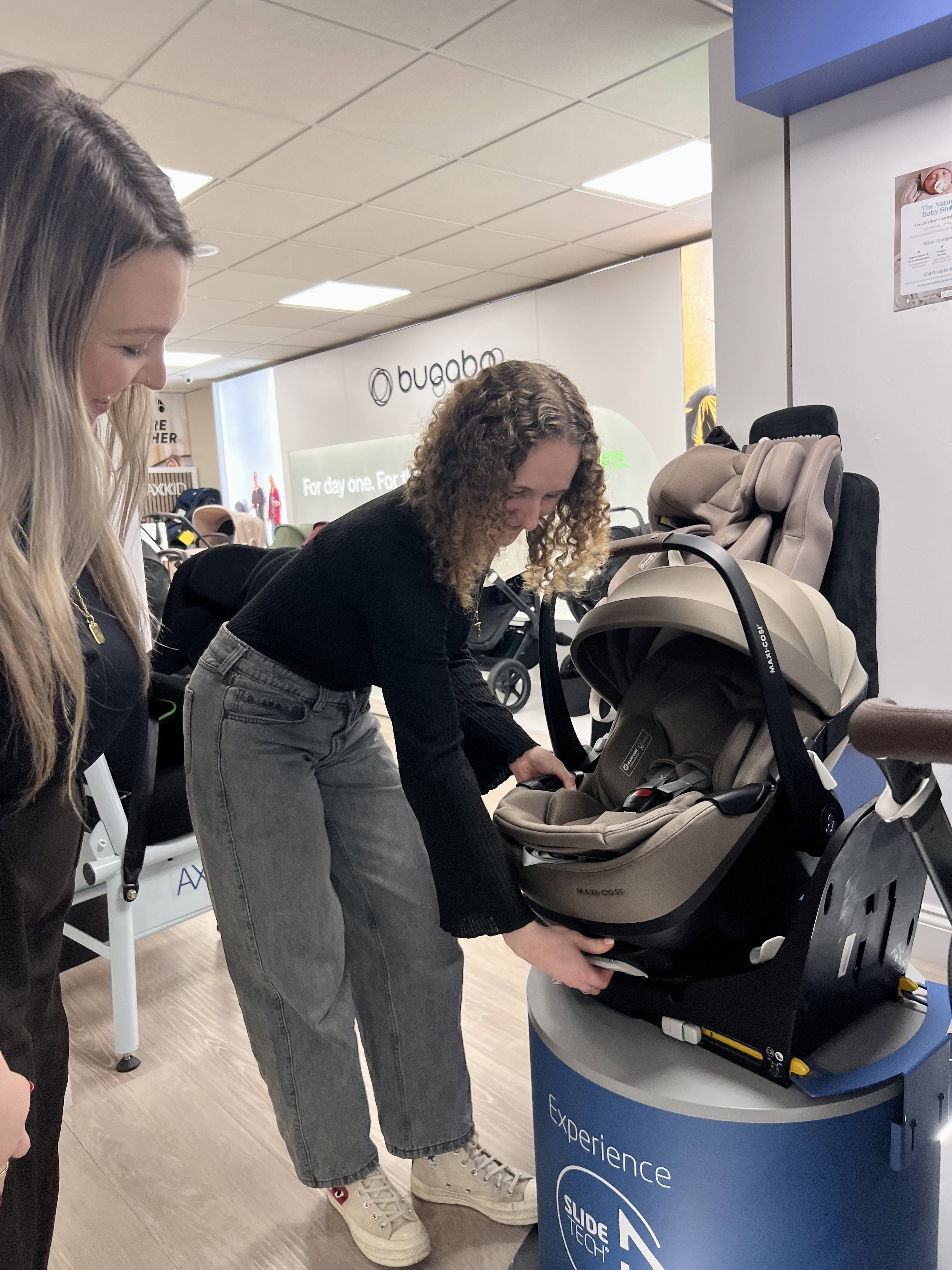 Nursery expert fitting an infant car seat onto an ISOFIX base in the back of a family car while an expectant mum watches