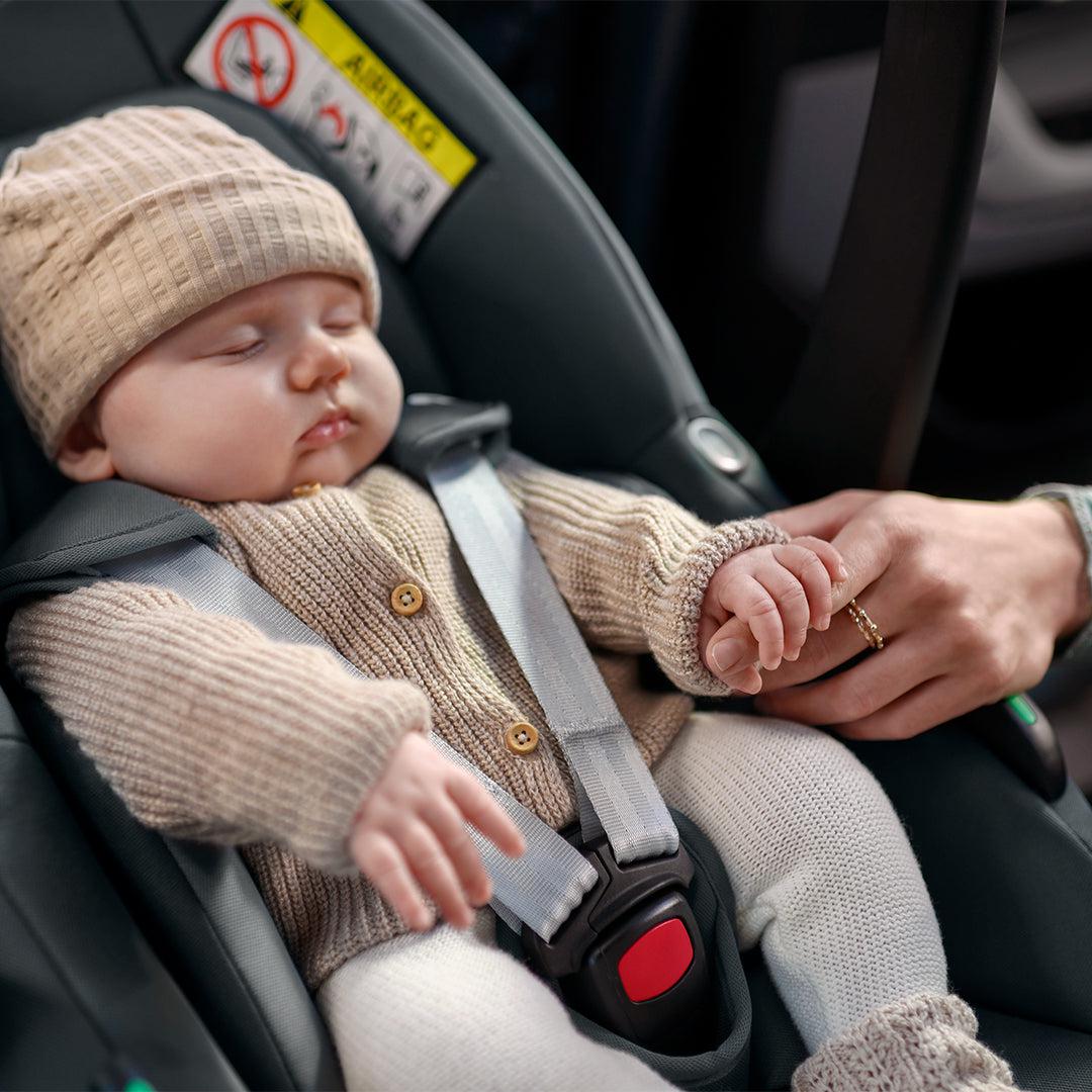 Smiling mother tucking a cream knitted blanket around her sleeping newborn in a taupe Silver Cross infant car seat on the back seat of a car