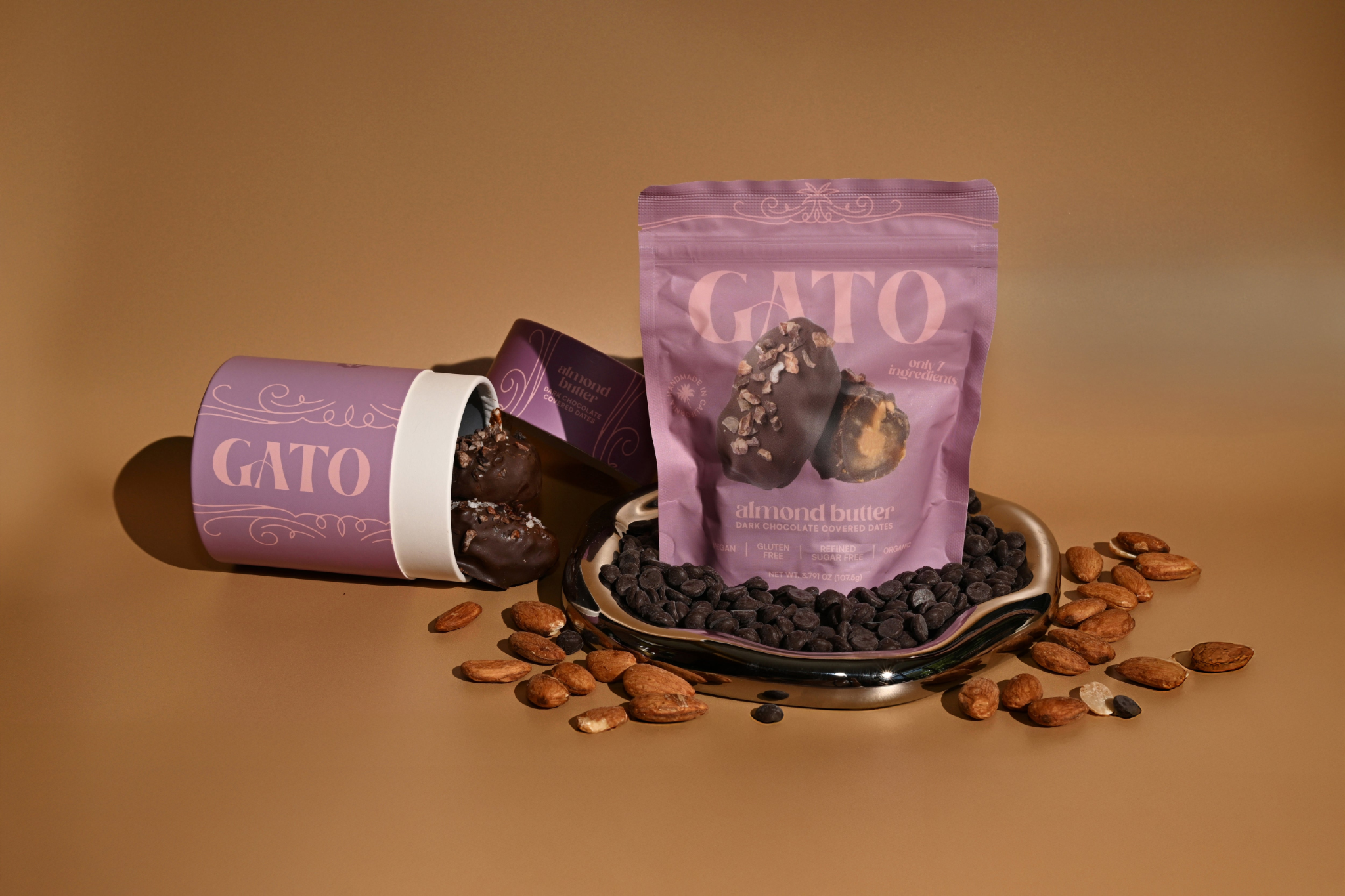 Gato dark chocolate covered dates in a lilac pouch and canister, surrounded by almonds and chocolate chips