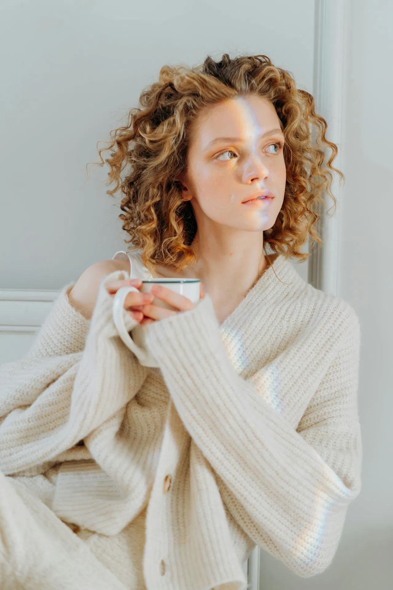 Frau mit lockigem Haar in einem gemütlichen cremefarbenen Pullover, die eine Tasse hält und nachdenklich zur Seite schaut.
