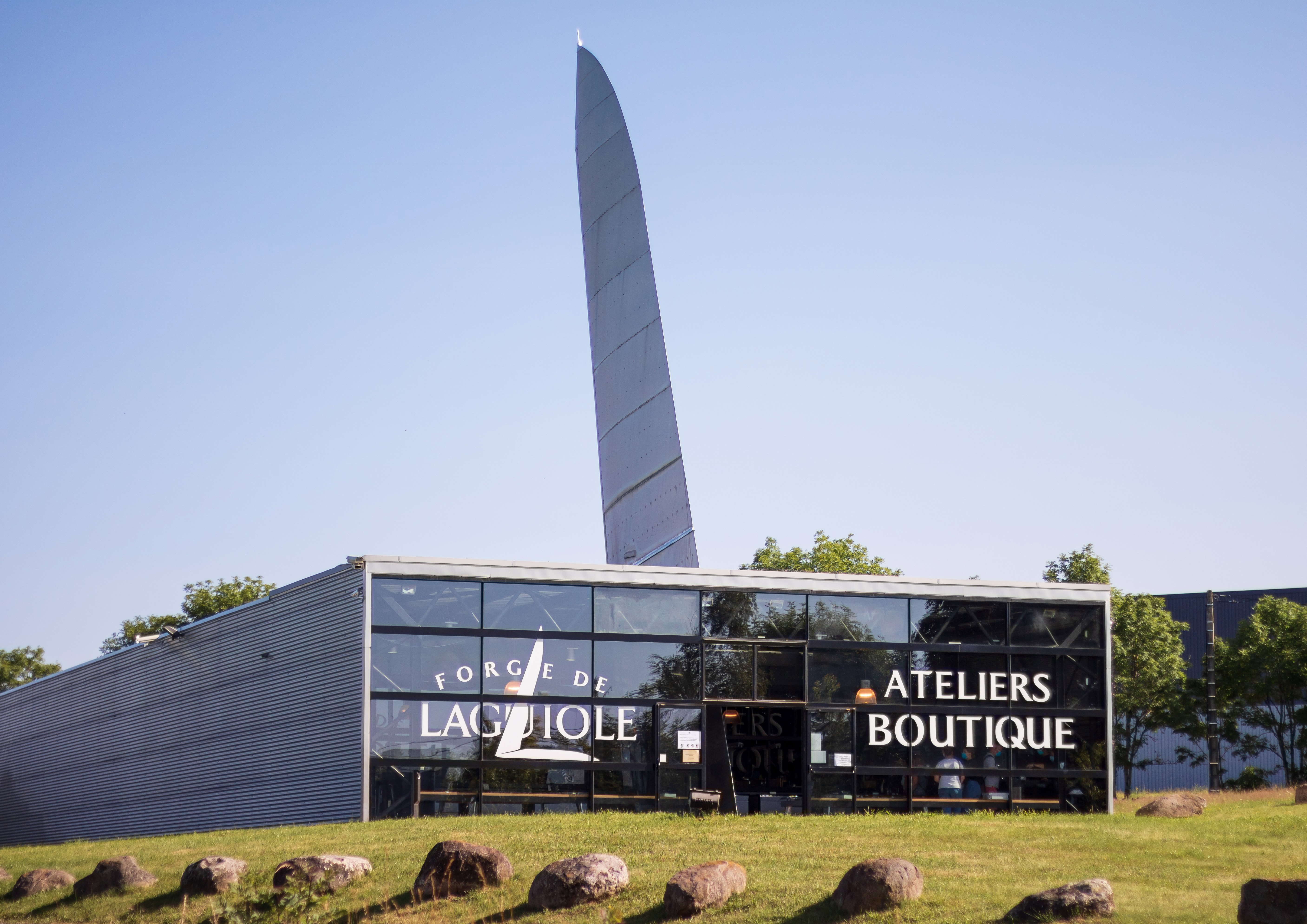 The Forge de Laguiole factory on the Aubrac plateau, its aluminium-clad workshop crowned by a giant steel knife blade, surrounded by green pastures and dry-stone walls