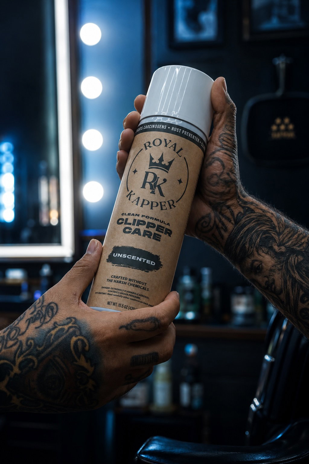 Tattooed barber holding a can of Royal Kapper Unscented Clipper Care spray in a dark barbershop