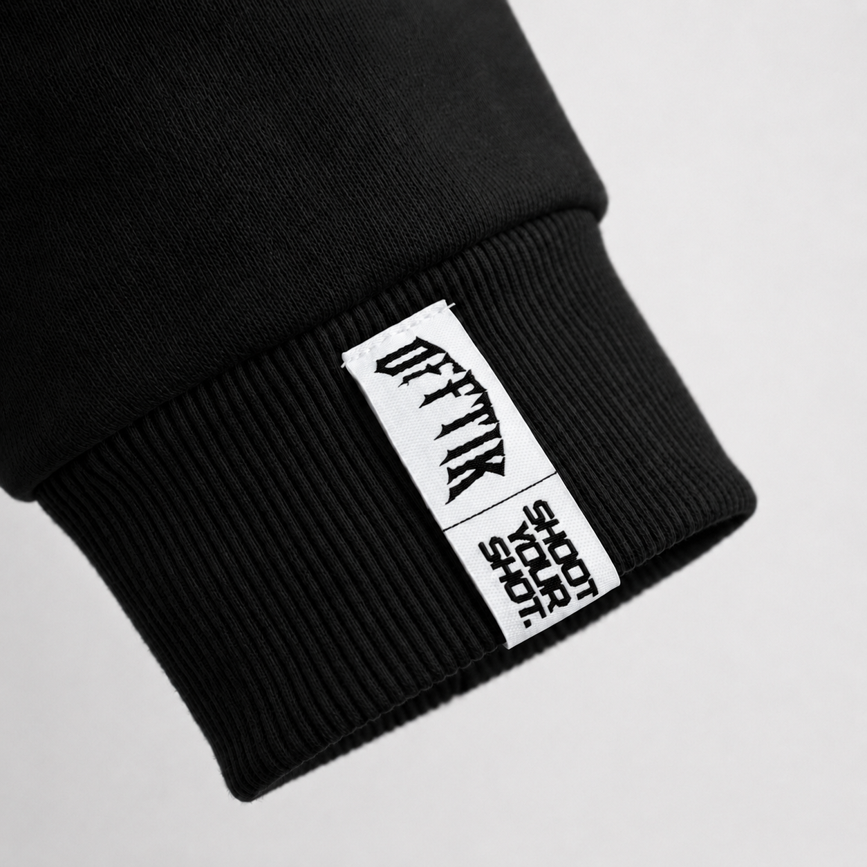 White woven OFFTIK Shoot Your Shot tag stitched onto the black ribbed cuff of the Throwy hoodie