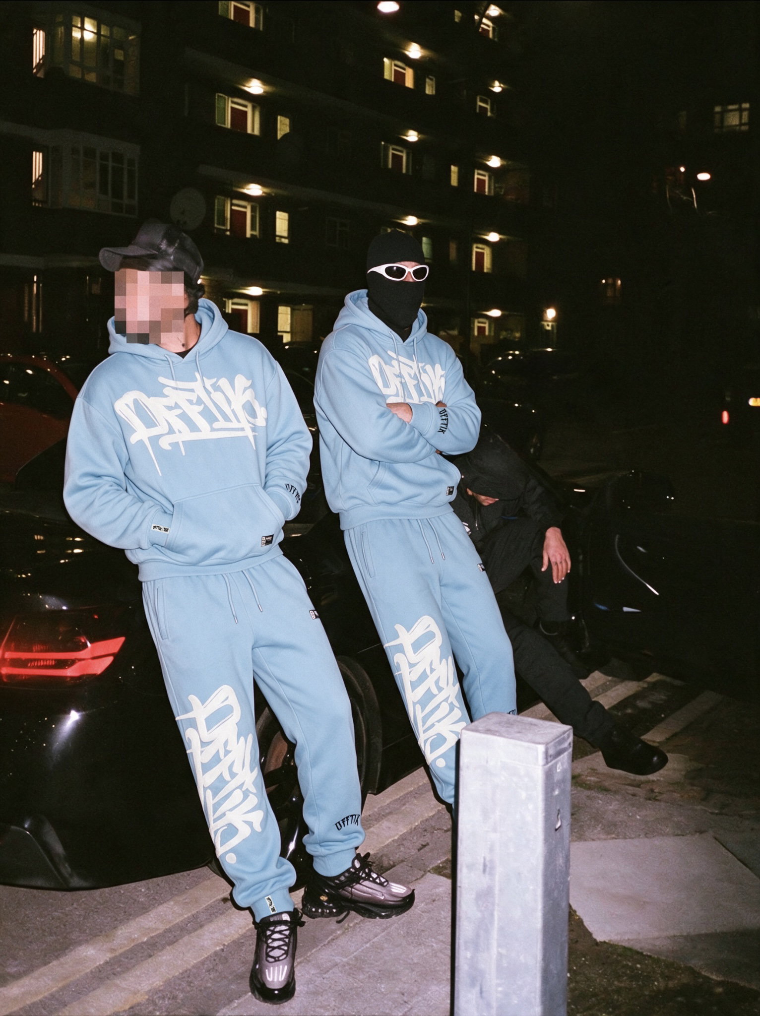 Two men in matching OFFTIK Signature Blue/White hoodies and trackies with cream chenille script logos, leaning on a black car outside a lit apartment block at night