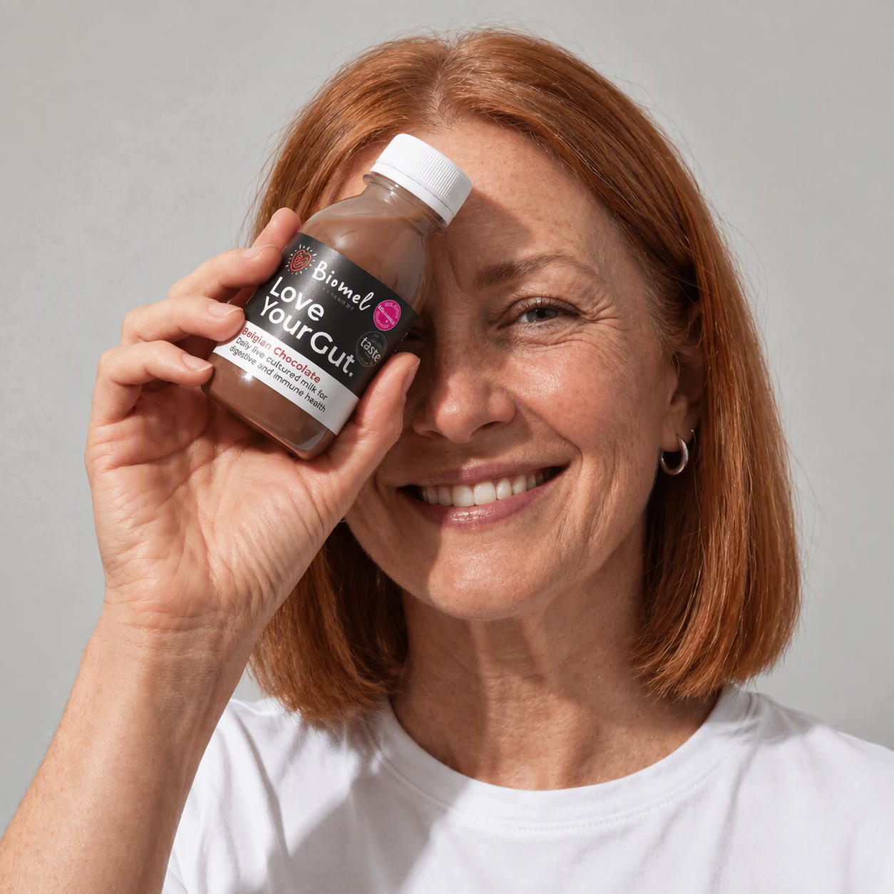 A middle-aged woman with red hair smiling and holding a brown probiotic drink bottle near her face