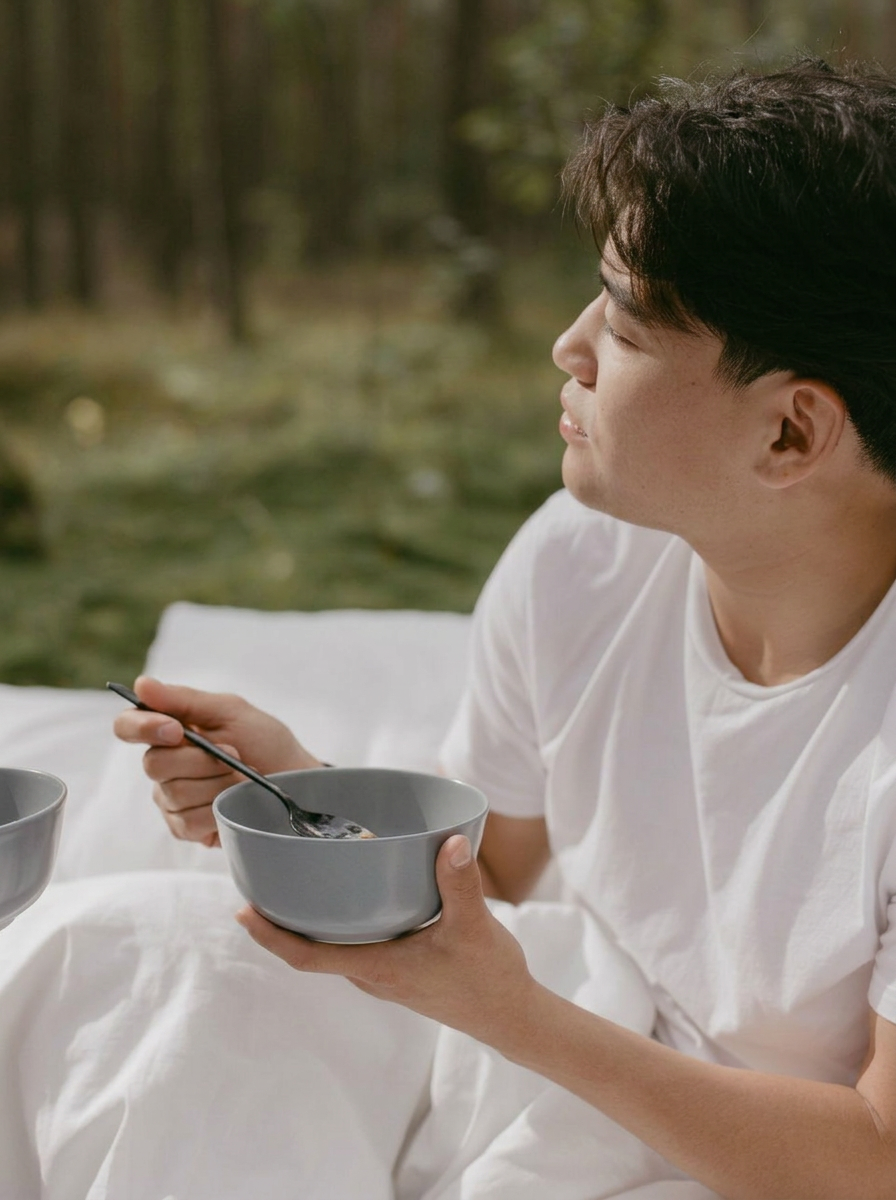 A man in a white shirt sitting outside holding a bowl and spoon, looking to the side.