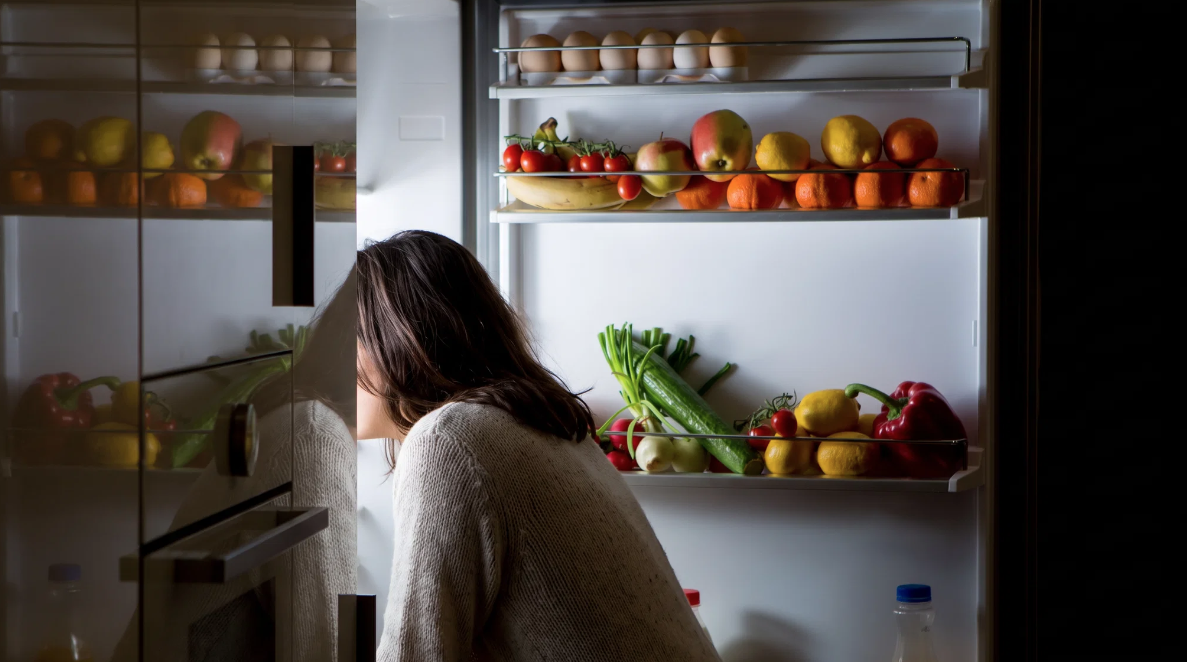 Mujer de espaldas mirando por la noche el interior de una nevera iluminada, con fruta y verdura en los estantes