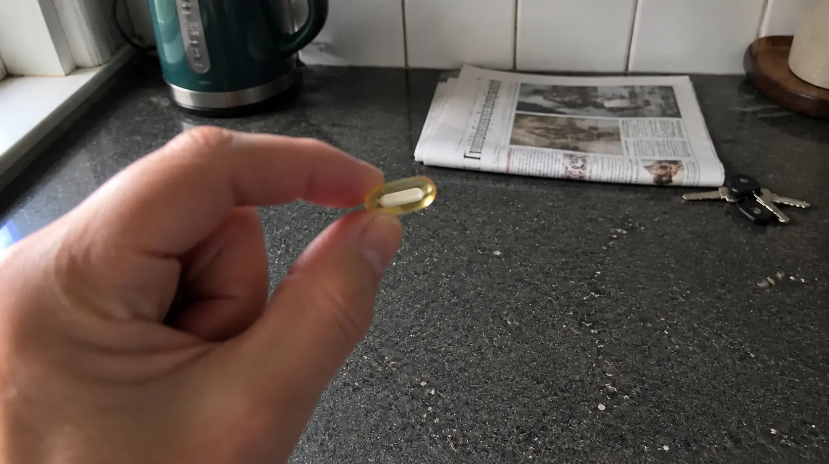 A golden Olivis cap-in-cap softgel held up to the light, showing the inner beadlet floating in olive oil