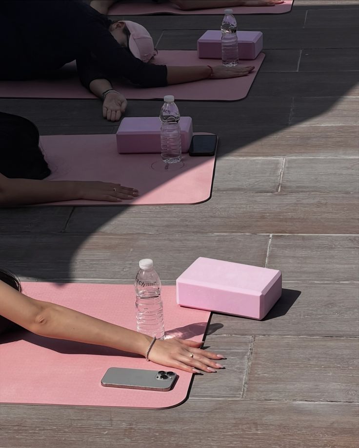People lying on pink yoga mats with water bottles, phones, and yoga blocks on a sunny deck.
