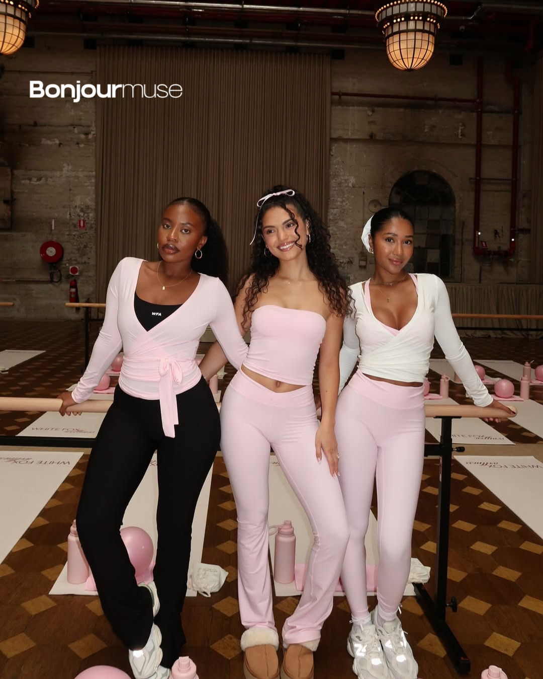 Three women in pink and black activewear pose in a studio with exercise mats and water bottles.