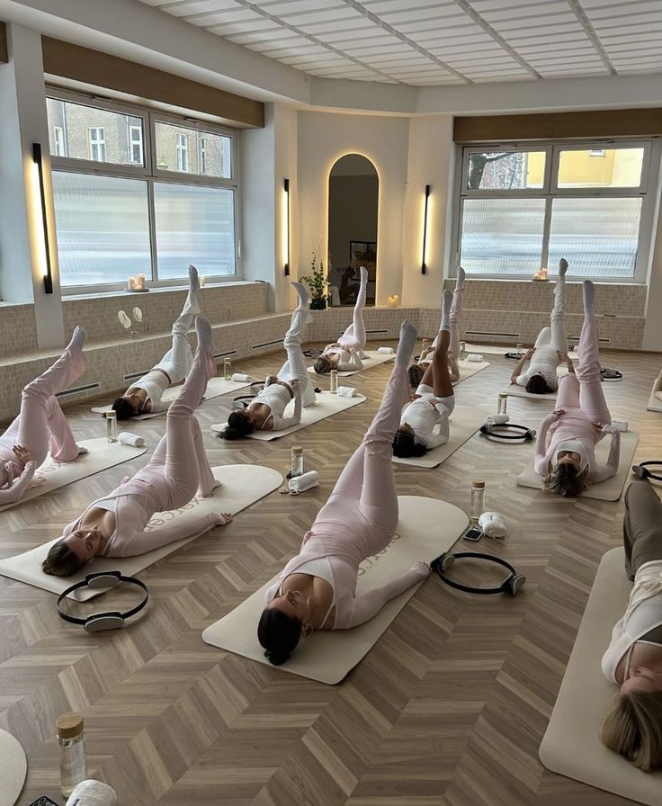 A group yoga class with participants in pink outfits, doing leg raises on mats in a bright studio.