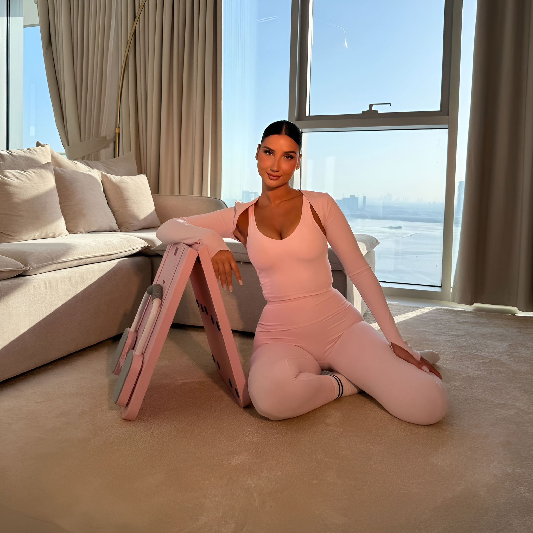 Woman in pink outfit kneeling by an exercise home pilates board in a sunlit room with large window views.