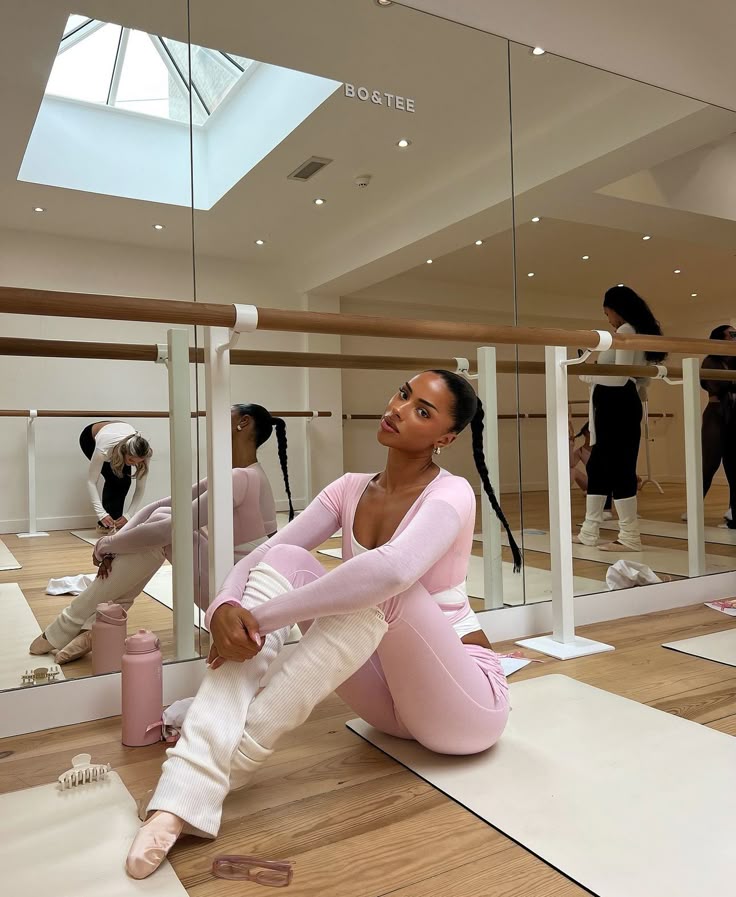 Woman in pink workout attire sitting on yoga mat in a studio with mirrors and ballet barre.
