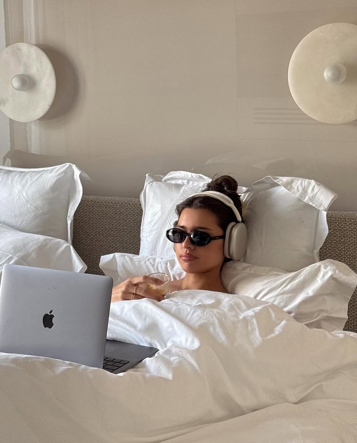 Person in bed with headphones, sunglasses, holding a drink, and using a laptop.