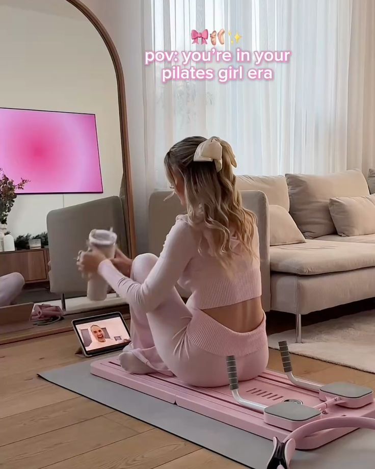 Woman in pink workout outfit on a Pilates mat, watching an exercise video on a tablet.