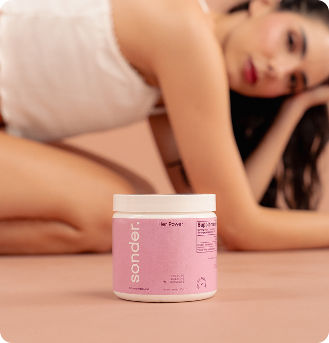 A pink container of Sonder “Her Power” 100% pure creatine monohydrate is placed in the foreground, with a woman in a white outfit kneeling and leaning to the side in the softly blurred background.