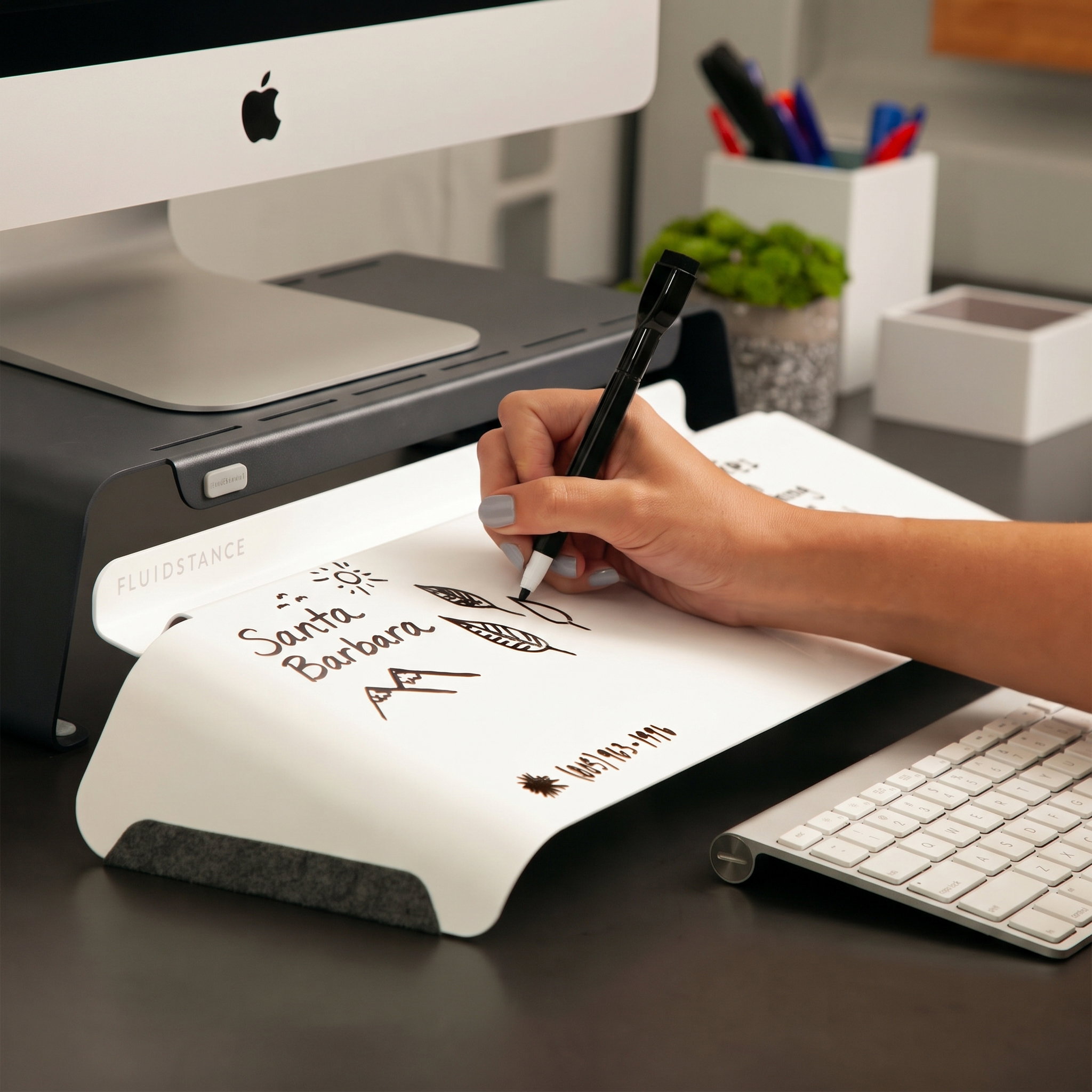 A hand writing notes on a white Slope desk whiteboard that sits between a keyboard and a monitor on a Raise stand