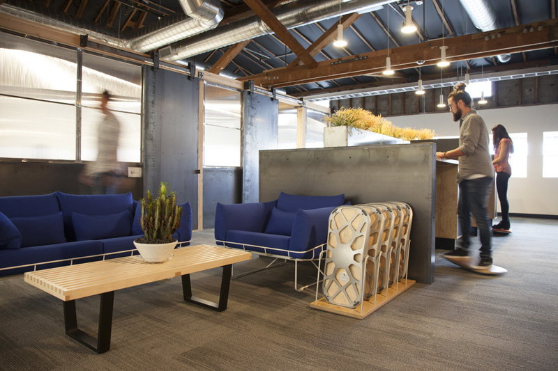An open industrial office lounge with blue sofas, a loaded rack of FluidStance decks on the floor and a colleague working at a standing counter on a deck