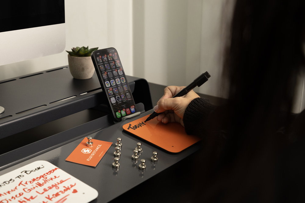 A person writing TODAY on an orange Flow Card stuck to a Slate Slope, with metal magnets, a handwritten white Flow Card and a phone docked in the Slope's rear channel