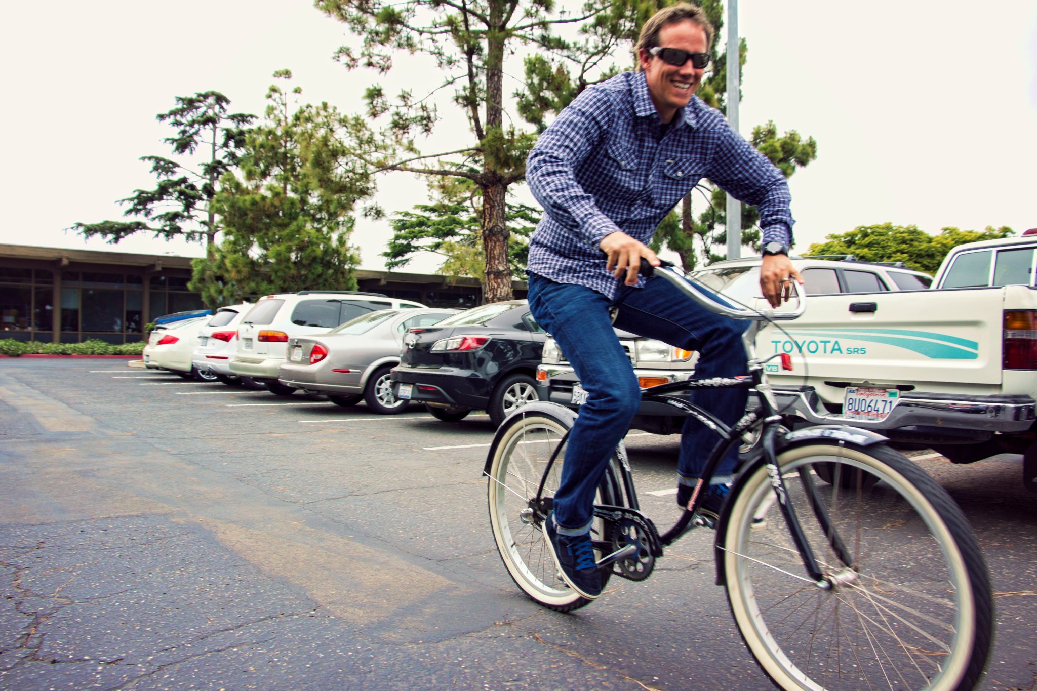 Smiling man in sunglasses and a plaid shirt riding a cruiser bike through a parking lot outside the office