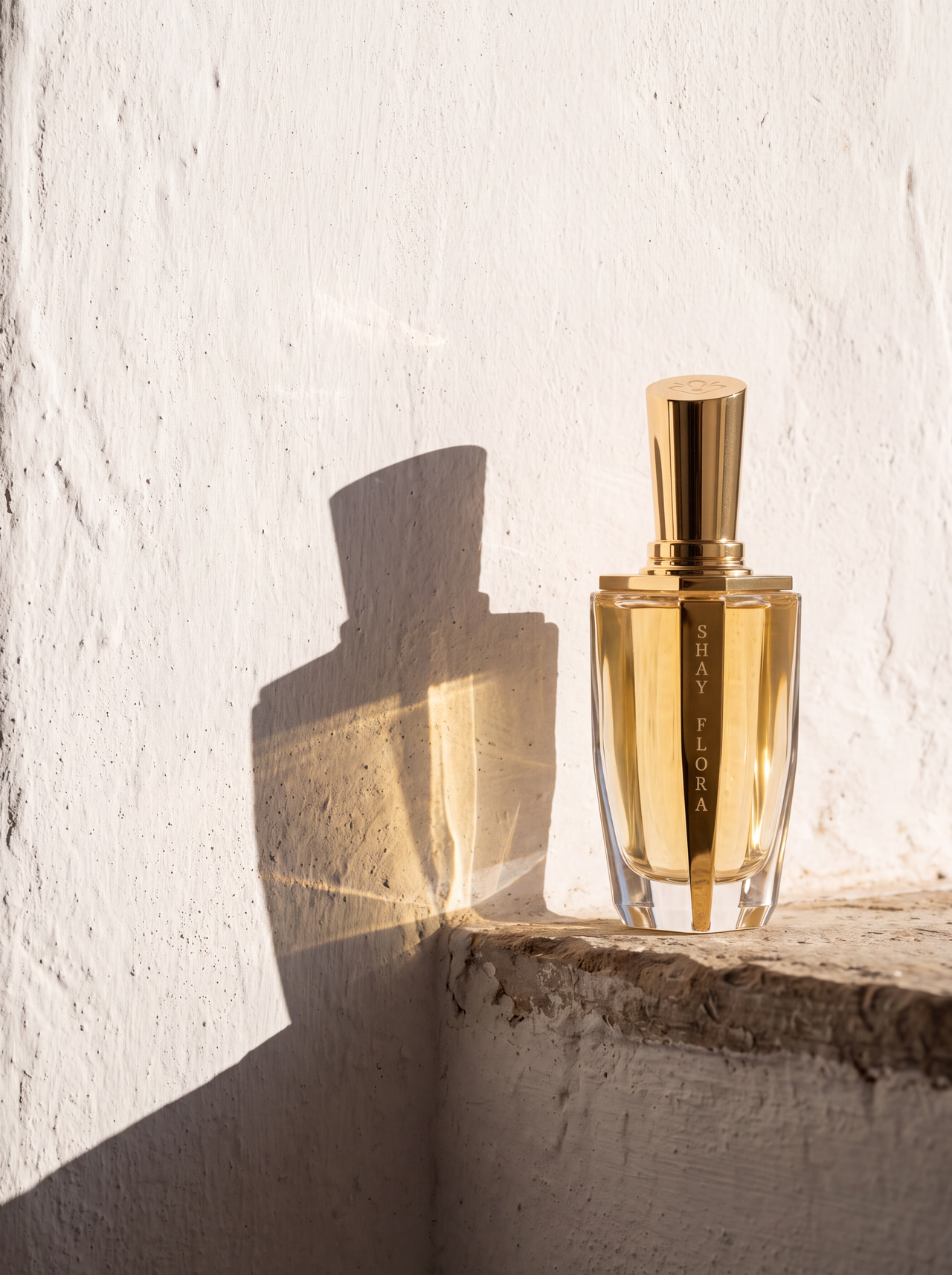 A golden perfume bottle labeled Shay Flora casts a shadow on a textured white wall
