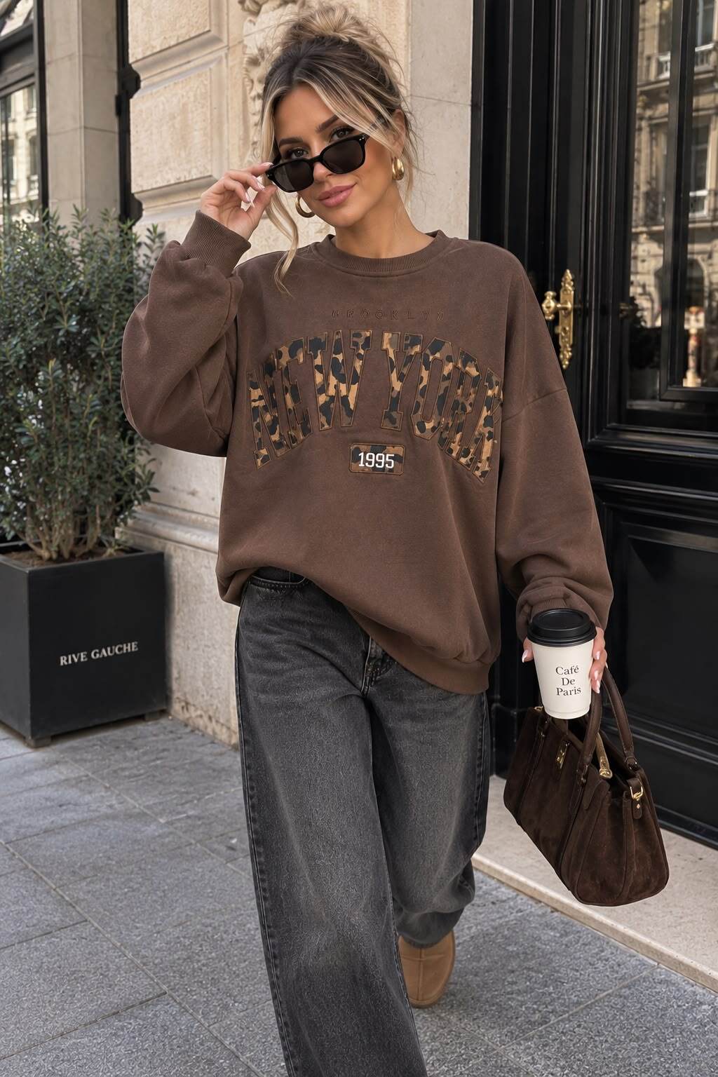 Frau im braunen Oversize-Sweatshirt mit New-York-Print, grauer Wide-Leg-Jeans und Coffee-to-go vor einer eleganten Stadtfassade