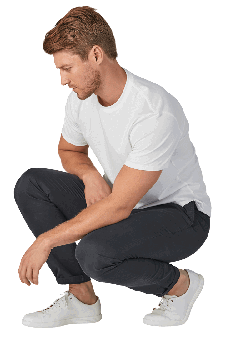 Man squatting, wearing a white t-shirt, dark chinos perk pants, and white sneakers, looking down.