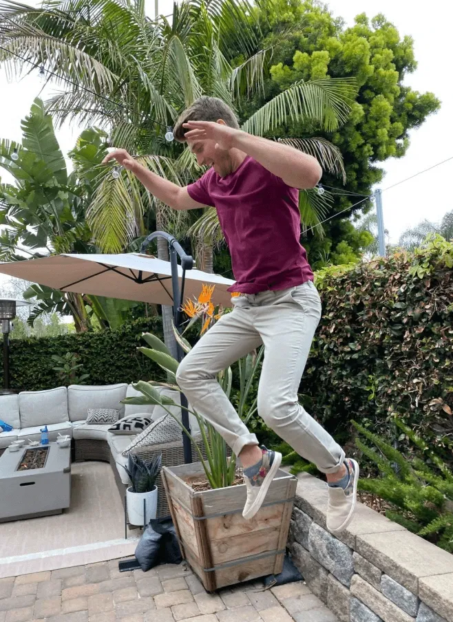 Man in maroon shirt jumping off a stone ledge in a garden with palm trees and patio furniture.