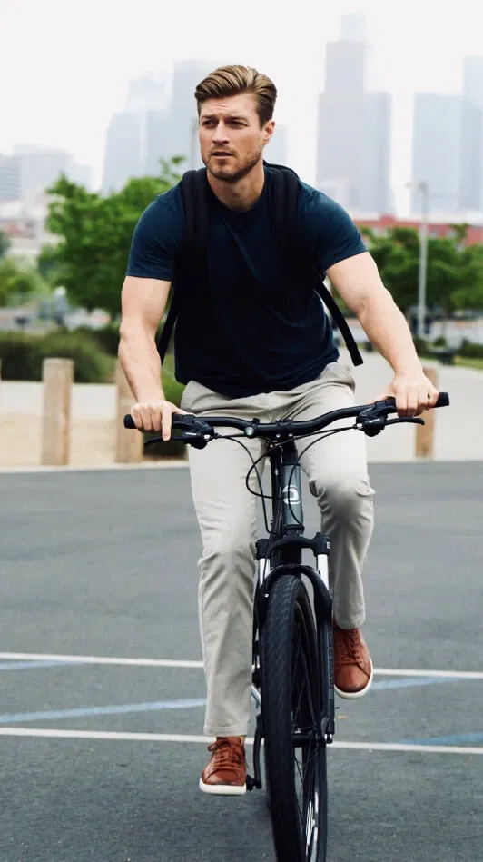 Person cycling on a road, wearing a navy shirt, khaki chino perk pants, and brown shoes. City skyline in background.