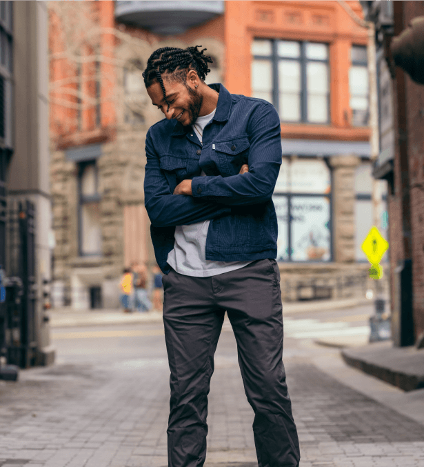 Man in denim jacket and wearing dark chinos Perk standing on city street with arms crossed, looking down and smiling.