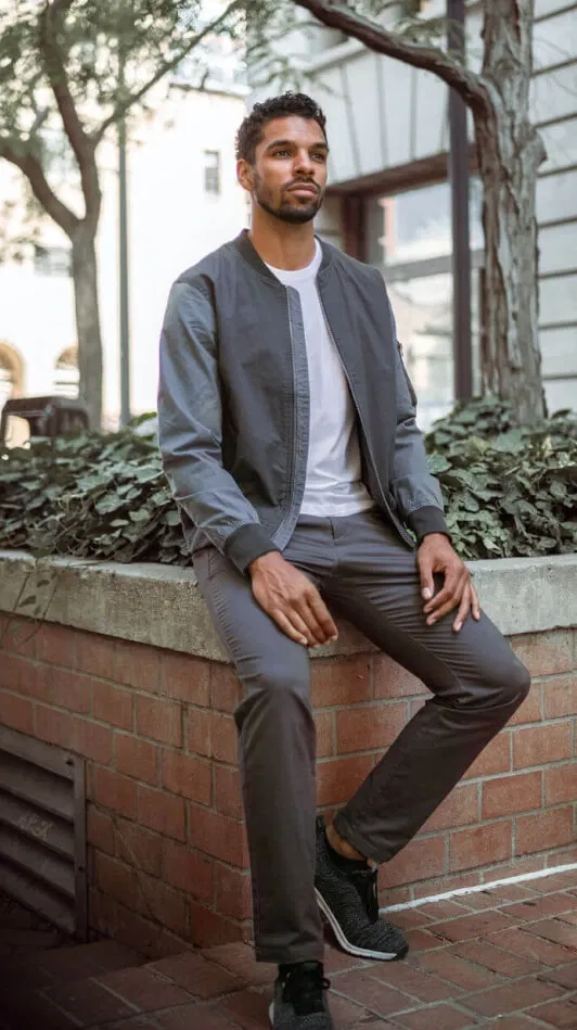 Man in gray jacket and perk chinos pants sitting on a brick planter, outdoors.