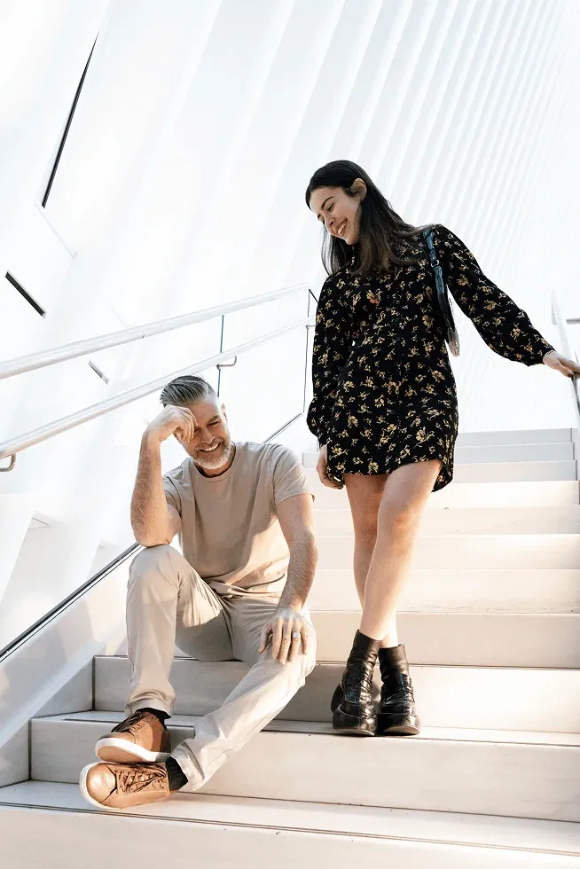 Man sitting on stairs, smiling up at woman standing in floral dress, inside modern white building.