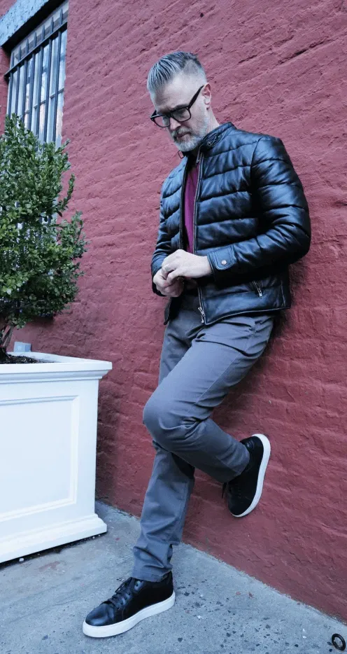 Man in black jacket, gray pants, leaning on red brick wall, looking down, hands clasped.