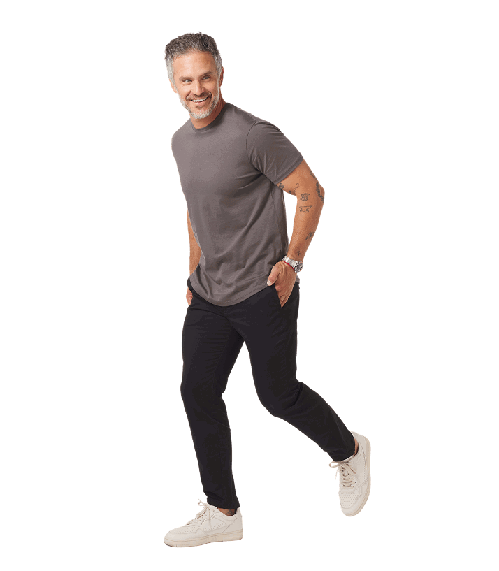 Man in gray shirt and dark pants smiling, walking forward with a hand in pocket, wearing white shoes.