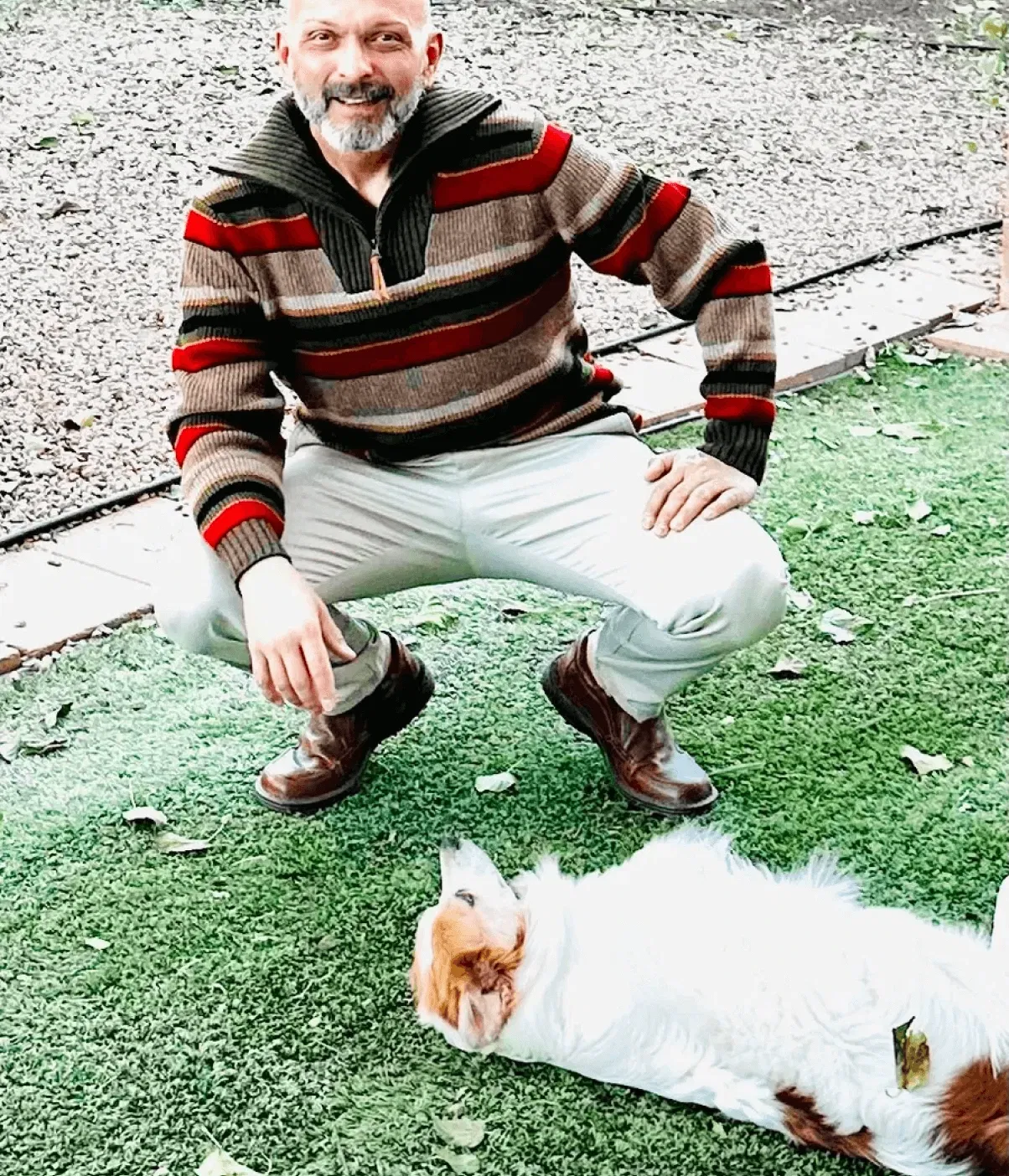 Man squatting on grass in a striped sweater, with a white and brown dog lying nearby.