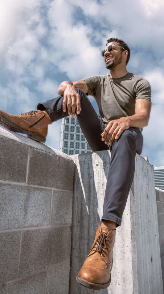 Man in sunglasses sits on concrete ledge, wearing brown boots and a gray shirt, with city building behind.