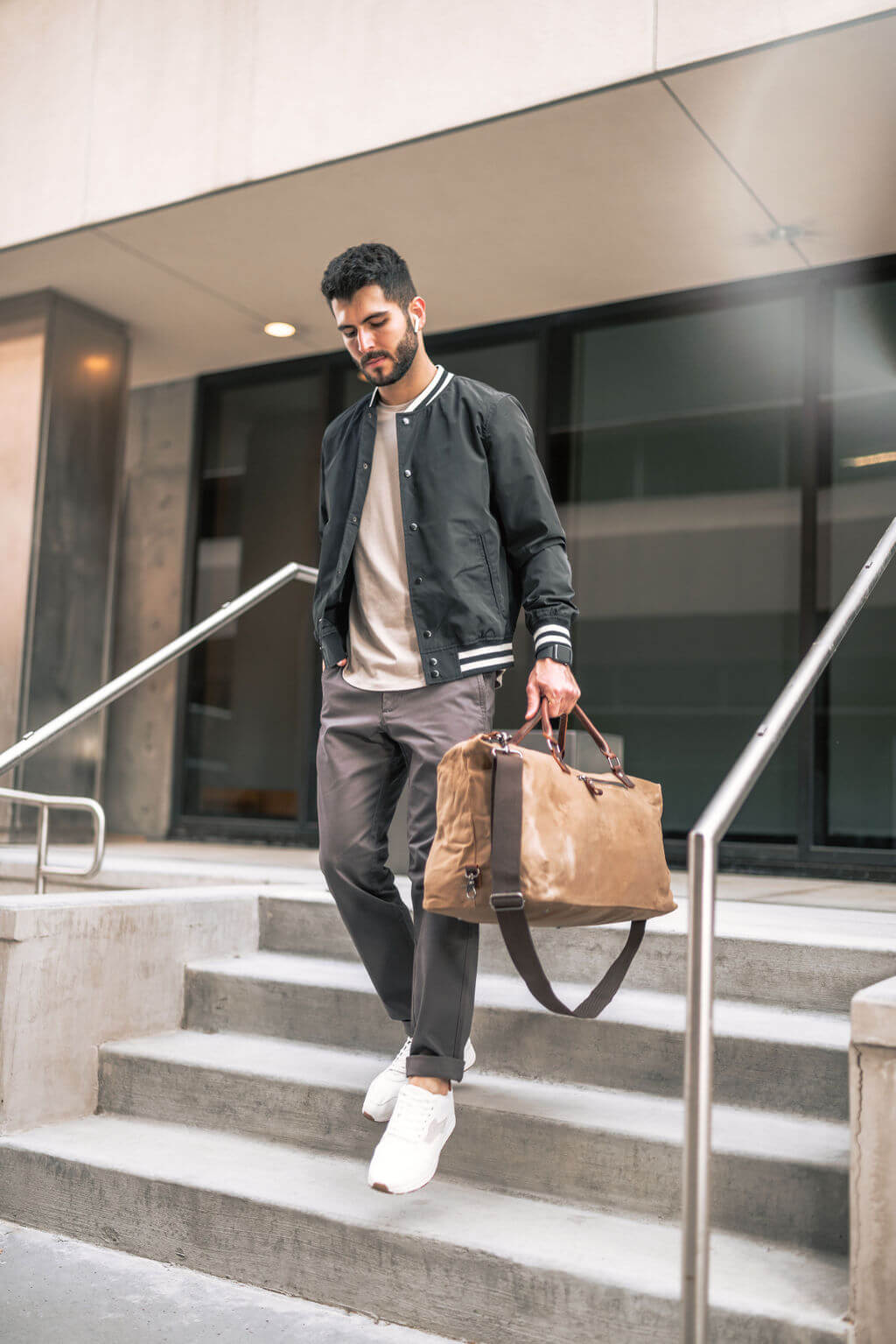 Man in casual outfit using a Perk Chinos descending stairs, holding a tan duffel bag, outside modern building.