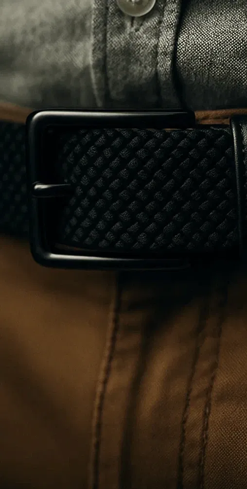 Close-up of a textured black belt with a shiny buckle on a gray shirt and brown pants.