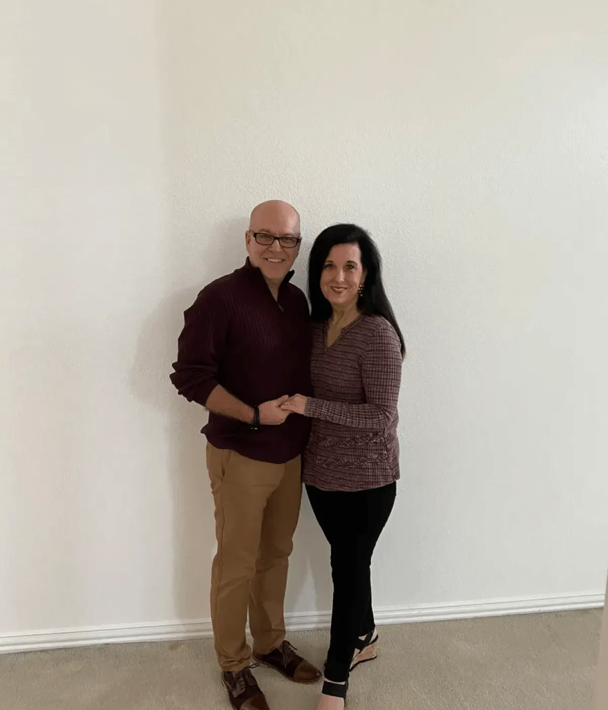 A couple stands smiling and holding hands against a plain white wall.