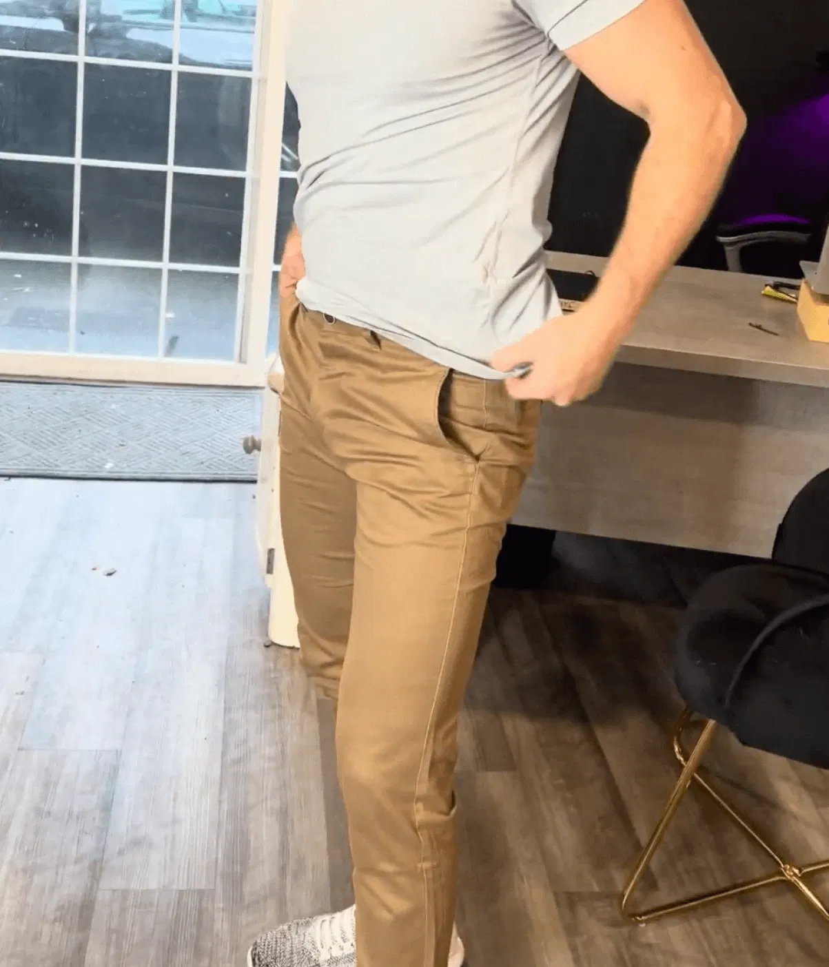 Person in a light gray shirt and brown pants stands beside a desk and black chair.