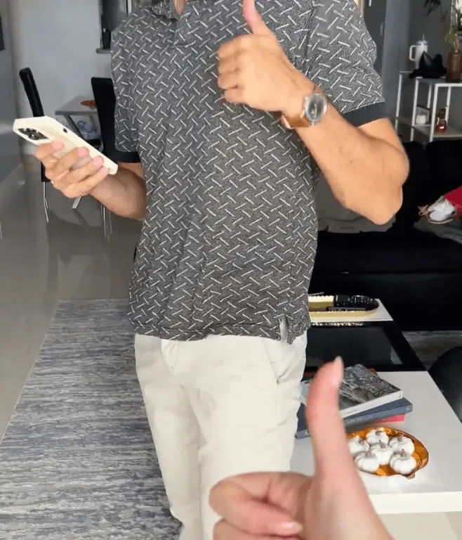 Person giving thumbs up, holding a phone, wearing a patterned shirt and light Perk pants in a living room.