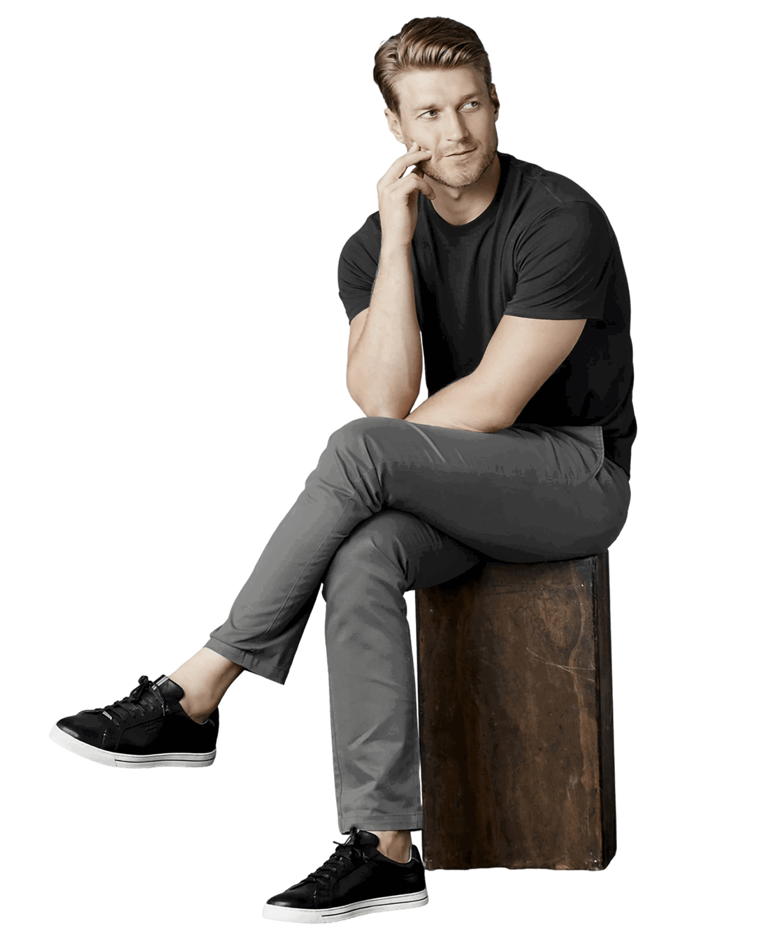 Man sitting on stool, wearing black t-shirt, gray pants and sneakers, looking thoughtful.
